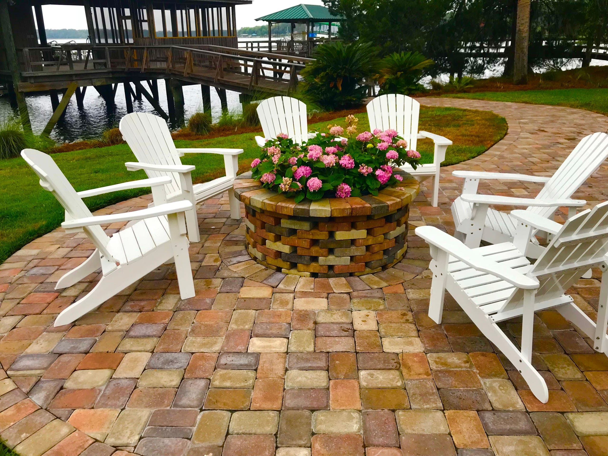 White Adirondack chairs circle a brick fire ring planted with hydrangeas, with the dock beyond.
