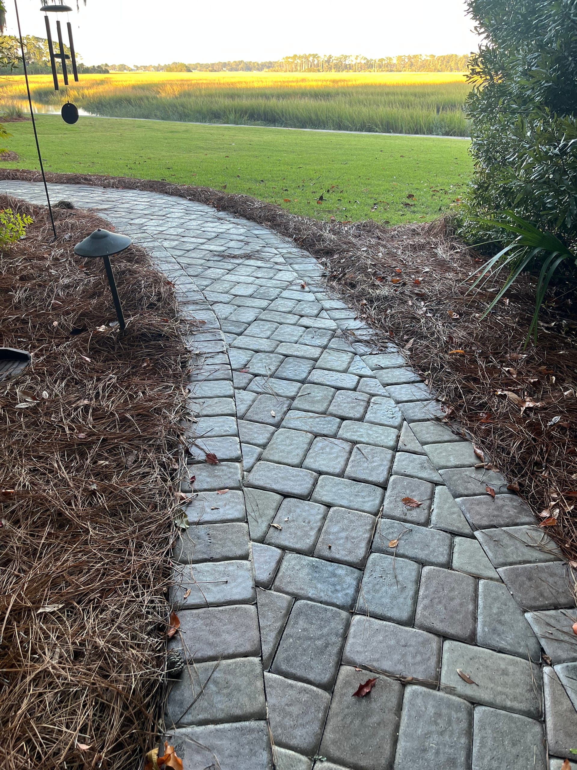 Paver path curving toward the open marsh, with a copper path light beside it in the pine straw.