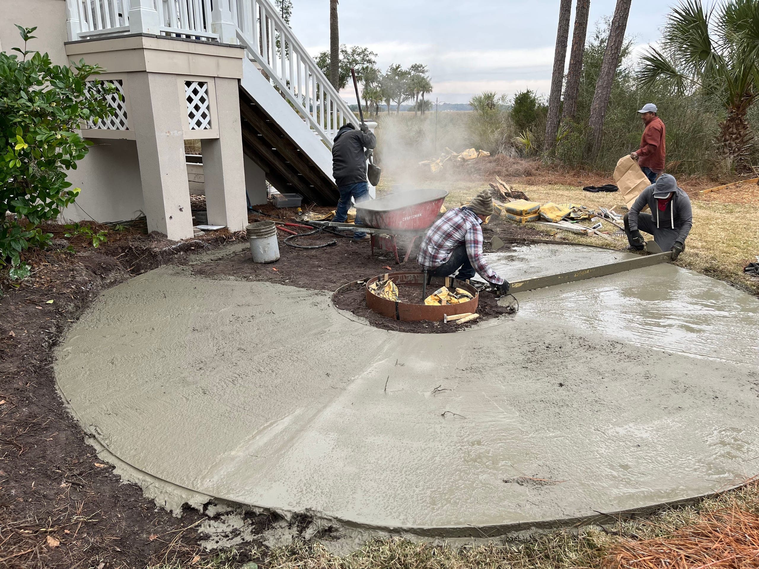 The crew screeds a fresh concrete pad at the marsh edge, the fire-pit ring formed at its center.