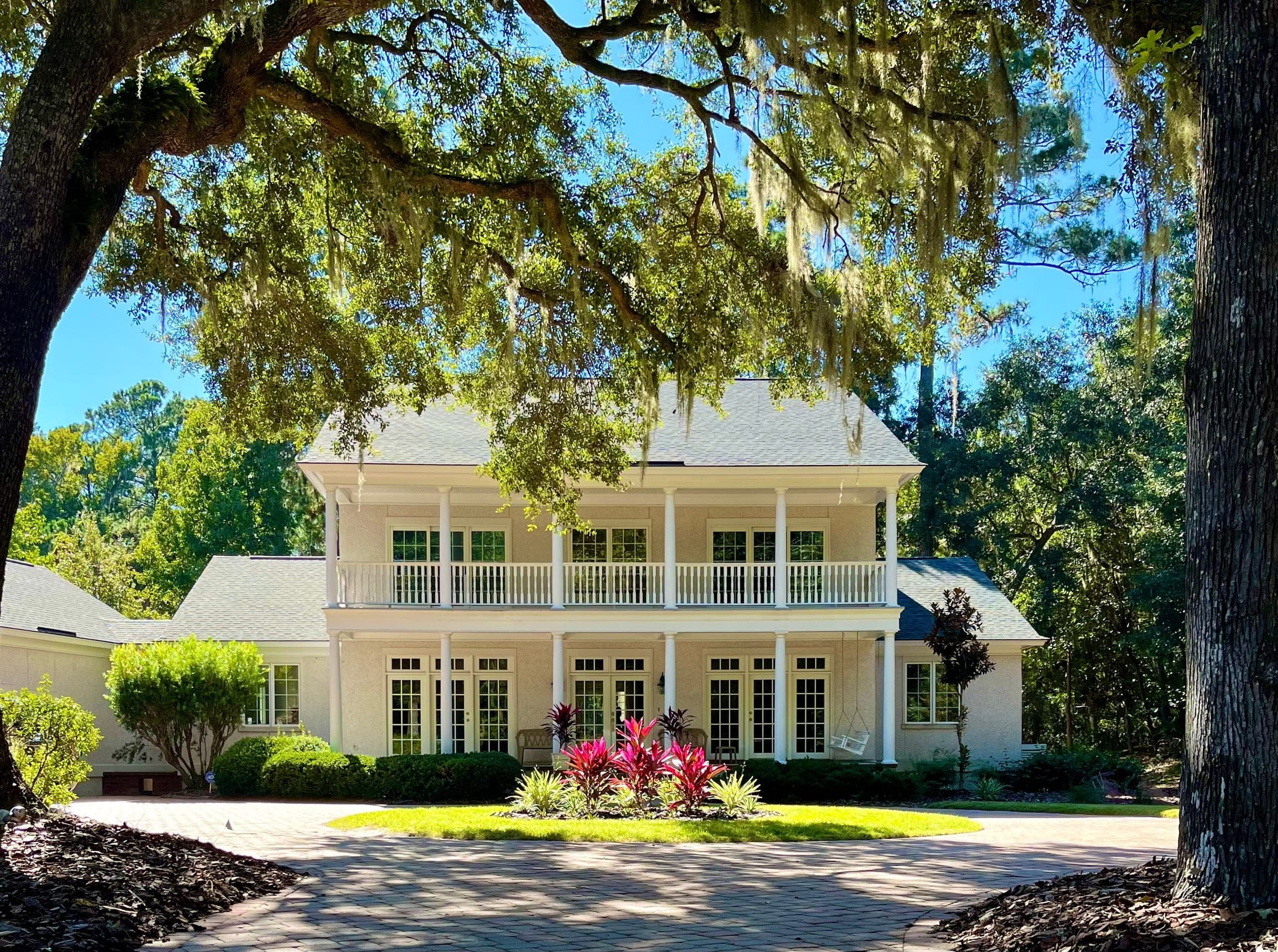 Live oaks frame a two-story house with double porches; a paver drive circles a planted island.