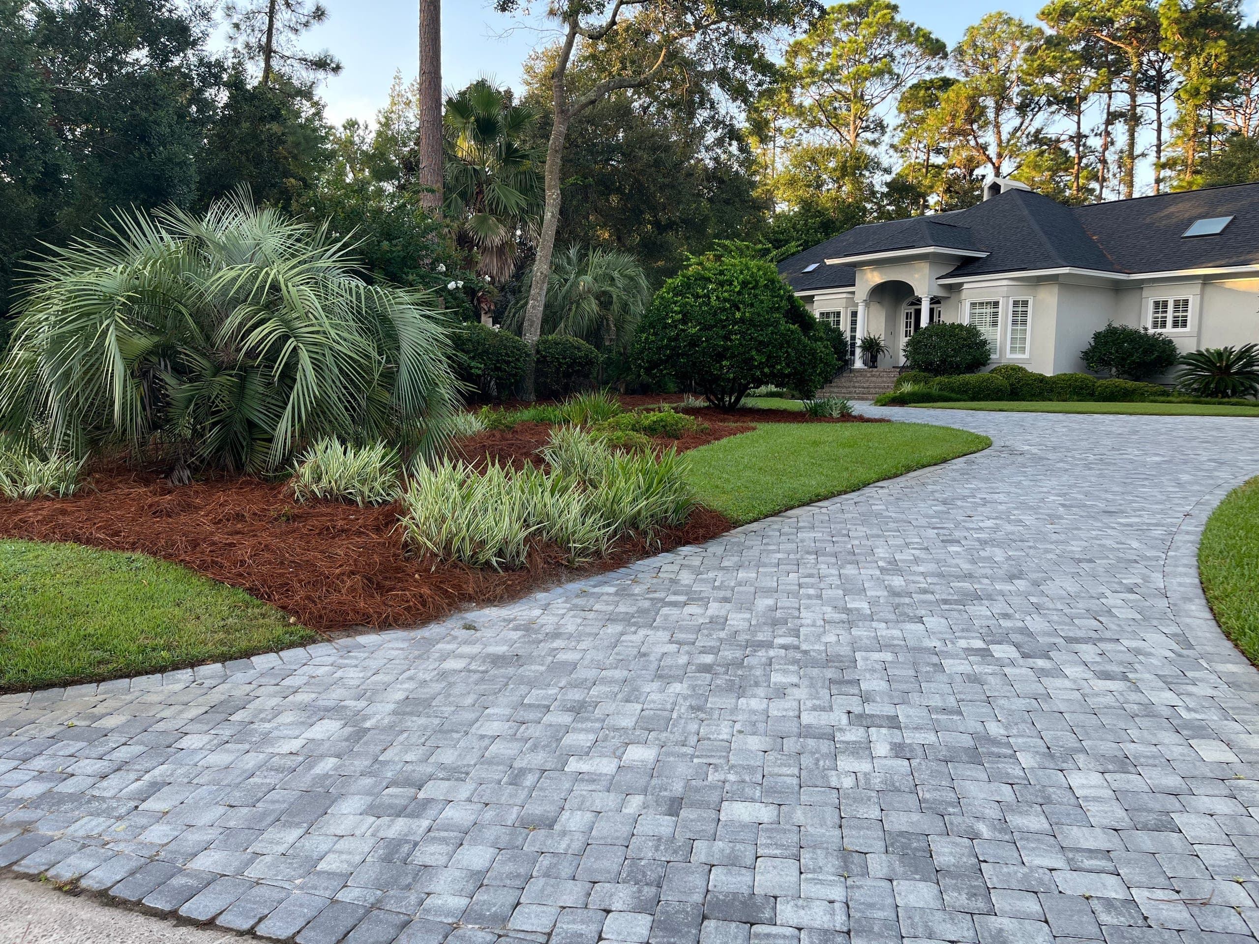 Tumbled paver driveway curving to the front entry between palms and planted beds.